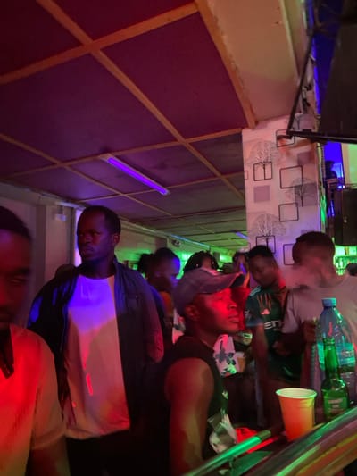 The image shows a bar in Dodoma, Tanzania (latitude: -6.163, longitude: 35.762). The bar has a wooden ceiling with red and purple lighting. There are multiple people in the bar, mostly men. The image captures the men in a dimly lit bar, with a focus on the individual in the foreground, who is wearing a blue cap, a black tank top, and a white floral print shirt. He is sitting at the bar, looking to the left with a thoughtful expression. There are other people in the background, some of whom are obscured by the light. There are plastic cups and drinks on the bar counter. The scene is a typical bar atmosphere, with people drinking and socializing. The image is taken from a low angle, which emphasizes the height of the bar and the people sitting at it. The focus is on the man in the foreground, who is the central figure in the image.