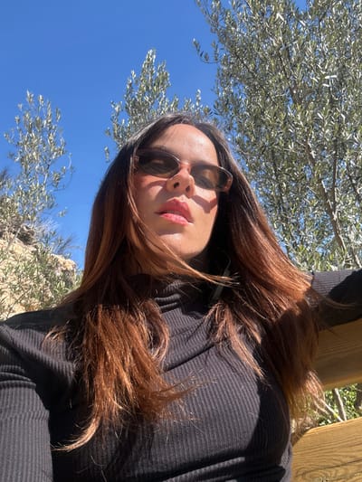 The image displays a close-up, low-angle outdoor portrait of a female individual under a clear, bright blue sky, consistent with the natural environment surrounding Alcoi/Alcoy, Spain. The woman, positioned centrally, has long, reddish-brown hair falling over her shoulders and chest. She wears dark-tinted, oval-shaped sunglasses and a black, ribbed, long-sleeved turtleneck top. Her lips are coated with a reflective, reddish-coral cosmetic. Strong sunlight illuminates her, creating distinct shadows on her face and upper body. The background features the top portions of several olive trees with silvery-green foliage, and a light-colored, rocky hillside is visible on the far left. A section of a light-colored wooden or composite structure, possibly a bench or railing, is visible at the bottom right corner of the frame.