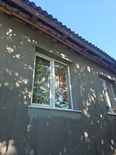 This is a low-angle, medium shot photograph of the exterior of a building under a clear blue sky. The building's wall is covered in gray plaster, with some areas showing a lighter, unfinished texture, suggesting renovation work. Sunlight casts dappled shadows of tree leaves across the facade. Two white-framed windows are visible on the wall. The left window is larger and reflects green foliage and parts of the sky. The right window is smaller and shows reflections of similar greenery and possibly some interior curtains. Above the wall, a section of the roof is visible, featuring wavy, dark gray roofing tiles supported by rough wooden beams. Insulation material, appearing light brown and fibrous, is exposed between some of the beams. The architecture suggests a residential building in Rădoaia, Moldova.