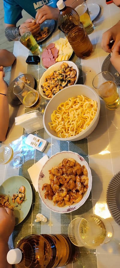 A group of people are gathered around a table laden with food and drinks. The scene appears to be a meal or gathering, possibly a party or family dinner. There are several dishes of food including shrimp, a cheese and meat platter, and a bowl of pasta or noodles. Drinks include glasses of beer and a large bottle of amber liquid, likely a beverage. The table is covered with a checkered tablecloth. One person is seen eating shrimp, while others appear to be engaged in conversation or preparing to eat. There is a small electronic device on the table, possibly a remote control or a timer. The lighting suggests an indoor setting, possibly a home dining area. The image is taken from a high angle, looking down at the table. Without further information, it is not possible to determine the specific location within the city of Dinskaya, Russia.