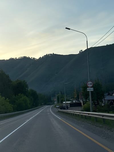 The image depicts a rural mountainous road in Manzherok, Russia, under an overcast sky transitioning from pale blue at the zenith to a faint yellow-white near the horizon, suggesting either early morning or late evening. A multi-lane asphalt road, marked with a solid double white line on the left and a solid yellow line separating the right lane from the shoulder, curves gently to the left into the distance. A metal guardrail runs along the right side of the road.
Dominating the background are steep, dark green mountains covered in dense coniferous forest, with a distinct tree line visible at the undulating crests. Several ridges are discernible, creating depth. In the midground, on the right side of the road, a black street lamp pole with a curved arm and a rectangular fixture stands prominently. Attached to this pole is a circular white road sign with a red border depicting two red cars facing each other, indicating a "no overtaking" or "no passing" zone. Below it, a white rectangular sign with an upward-pointing arrow states "200M," signifying the restriction's length.
Numerous power lines run horizontally and diagonally from the street lamp pole and other poles further down the road. To the right of the road, behind the guardrail, dense green foliage partially obscures glimpses of residential structures, likely wooden houses, indicative of a village or settlement. A white car, possibly a sedan, is parked on the right shoulder further down the road. Near some obscured buildings, a dark-colored utility vehicle or ATV is parked, with the Cyrillic word "ОКАТ" visible on a building facade. No people are visible in the scene.