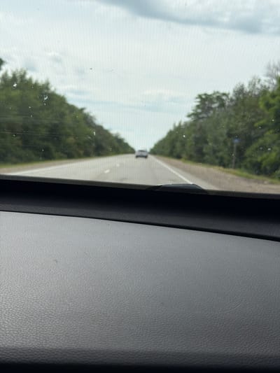 The image shows a view from inside a car looking forward through the windshield. A grey, textured dashboard is in the foreground. The car is on a paved road with a white line marking the lane. Trees line both sides of the road, and the sky is overcast. In the distance, another car is visible on the road. There is no specific information in the image to identify the location as Yugo-Severnaya, Russia.