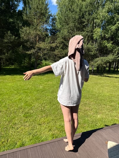 An outdoor, sunlit scene in Копнино, Russia, depicts one individual from behind, standing barefoot on a dark brown slatted wooden deck. The person, whose gender is not explicitly identifiable but appears to be female, is wearing a loose-fitting white t-shirt that extends to mid-thigh. A light brown towel is draped over their head and shoulders, partially obscuring the head. Their right arm is extended straight out to the side, palm open and facing forward, with fingers slightly spread and nails painted white. A distinct shadow of the person is cast on the deck, indicating bright sunlight from the left. Beyond the deck, a large expanse of vibrant green grass, interspersed with small yellow flowers, fills the midground. The background consists of a dense forest line dominated by tall pine trees and some lighter-barked deciduous trees. Patches of clear blue sky with minimal white clouds are visible through the tree canopy. A small corner of a light-colored cushion or mattress is visible on the far right of the wooden deck.