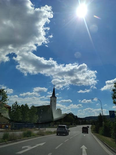 This is a photograph taken from a moving vehicle, looking out through the windshield. The scene depicts a road in Strakonice, Czechia, on a sunny day with scattered white clouds in a bright blue sky. The sun is high and bright, creating lens flares and starburst effects in the image.
In the foreground, a two-lane asphalt road is visible, with white lane markings. The vehicle from which the photo is taken is traveling on the right lane, with a white arrow painted on the road indicating a choice of going straight or turning right. Two cars are ahead: a white SUV in the right lane and a dark red minivan in the left lane. Further down the road, a smaller, lighter-colored car is visible in the distance.
To the left of the road, there are some buildings and trees. A prominent white church with a dark spire and a cross on top stands against the sky, surrounded by green trees. In front of the church, there are some temporary blue structures and yellow bins.
To the right of the road, there is a sidewalk or verge with vegetation, followed by trees and then a blue parking sign indicating a parking area 200 meters ahead. Beyond the immediate roadside, the landscape appears to be hilly with more trees and buildings visible in the distance. The overall impression is a suburban or semi-rural setting.
