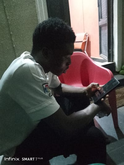 A dark-haired young man sits looking at a phone, which he holds in his hands. He wears a white collared shirt with a crest on the sleeve. The man sits in a red plastic chair. Behind him, another chair with a different design is partially visible. There's a window with bars over it, and the walls are a light color. The lighting is dim. The words "Infinix SMART 7 PLUS" are in the bottom left corner.