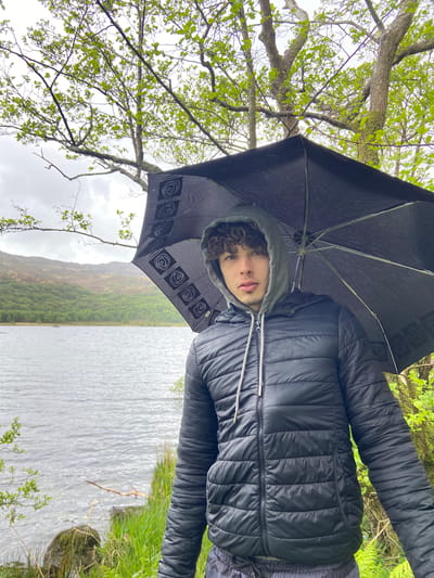 A person stands under a black umbrella with a spiral pattern on its edges. The individual is wearing a black hooded jacket. Behind the person, a body of water stretches toward a hillside covered in trees. Grass grows on the water's edge in the foreground. A tree branch with green leaves is also visible. The location appears to be outdoors, possibly a lake or reservoir in a natural environment. Given the city mentioned, the location is likely in or near Beddgelert, United Kingdom.