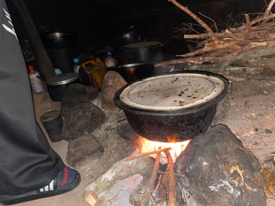 In the city of Junyura, Nigeria, a nighttime scene unfolds around a crackling campfire. A large, dark pot with a white lid sits precariously on rough-hewn stones, directly over the flickering flames. To the left, the lower portion of a person's body is visible, clad in black pants and athletic shoes with red and blue striped socks. The surrounding environment is cluttered with various items, including a yellow plastic jerrycan, a silver pot with a lid, a black pot, and a tall, dark cylindrical object. Several other pots and a pile of dry branches are also present, suggesting preparation for cooking or a recent meal. The ground appears to be dirt and stone, illuminated by the fire's glow.