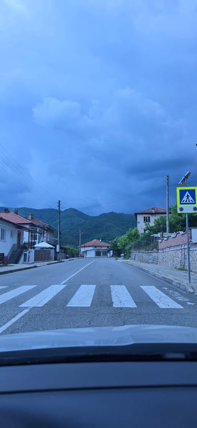 The image depicts a street scene in the village of Taran, Bulgaria, under a dramatic, dark, cloudy sky. A pedestrian crossing with white stripes is prominently featured in the foreground, painted across an asphalt road. To the right of the road, a yellow pedestrian crossing sign with a blue silhouette of a person is visible on a pole, illuminated by a solar panel. Beyond the crossing, several buildings with tiled roofs are visible, along with trees and lush green mountains in the background. Power lines stretch across the sky, adding to the somewhat somber atmosphere, likely due to the impending weather. The view appears to be from inside a vehicle, as the lower part of the image shows the interior of a car. There are no people or animals clearly visible in the scene. The overall impression is one of a quiet, possibly rural or semi-rural, setting on the verge of a weather event.