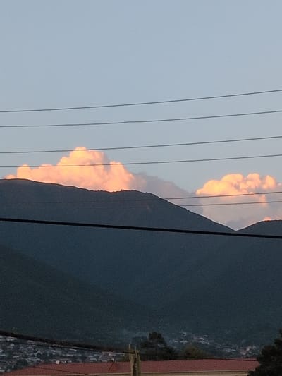 This is a vertically oriented, outdoor, low-angle photograph of a mountainous landscape at dusk. The scene is divided into two main sections by a dark, silhouetted mountain range that runs diagonally from the upper right to the lower left of the frame.

The left side of the image is dominated by a pale, twilight sky, traversed by three thin, dark power lines. Against this sky, a vibrant, orange-pink cloud formation is visible, brightly illuminated by the setting sun. The clouds appear to be cumulus, with a textured, billowy appearance.

The right side of the image is filled with the dark mass of the mountain, which appears to be covered in dense vegetation, suggesting a natural, undeveloped area. Along the very edge of the right side, there are hints of structures, possibly buildings, with some red and patterned elements visible, indicating the presence of a nearby settlement.

The time of day is clearly dusk, as indicated by the low angle of the light and the color of the sky and clouds. The weather appears clear, apart from the illuminated clouds. The location cues suggest a mountainous region, possibly with a town or village nestled in or near the mountains. No people or specific objects are clearly discernible, apart from the power lines and the distant structures. The overall mood is one of serene natural beauty, enhanced by the dramatic colors of the sunset. There is no visible text in the image.
