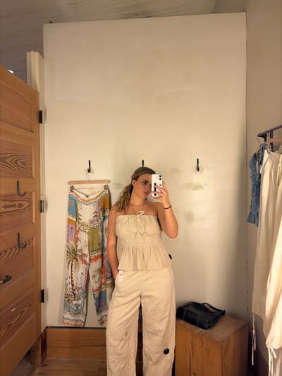 A woman stands inside a dressing room, holding a smartphone to take a selfie in the mirror. She's wearing a light-colored, two-piece outfit. Hanging on the wall is a colorful garment. The dressing room has a wooden door and is furnished with a wooden bench. A black bag is placed on top of the wooden bench. The specific location in Coral Gables, United States of America cannot be determined from the image alone.
