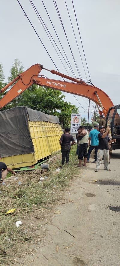 The media file captures an outdoor scene in Peurupok, Indonesia, featuring an excavator and a truck. The primary subjects are several men gathered around the excavator and a partially overturned yellow truck covered with a black tarp.
The excavator, an orange Hitachi, is positioned with its boom extended over the truck, suggesting an attempt to lift or move it. The truck appears to be off the road, partially in a ditch filled with mud and debris, indicating a possible accident or recovery operation. The surrounding area is somewhat overgrown with grass and vegetation. A paved road is visible to the right, with some scattered litter on it. In the background, there are trees and utility poles with many hanging wires overhead.
The people are dressed in casual clothing. Their postures and proximity suggest they are involved in the situation, some observing and others potentially directing the operation. One person is holding a phone, possibly taking a picture or video. The general atmosphere seems to be one of concern or focused attention on the task at hand.
A visible sign in the middle ground reads "COFFEE SHOP" at the top, followed by "KUPI ABUNAWAL" in larger text, indicating a local establishment. The lighting suggests it is daytime, with overcast or bright, diffused light. The weather appears to be warm and possibly humid, typical of the tropical climate. The overall scene depicts a challenging situation involving heavy machinery and a disabled vehicle.