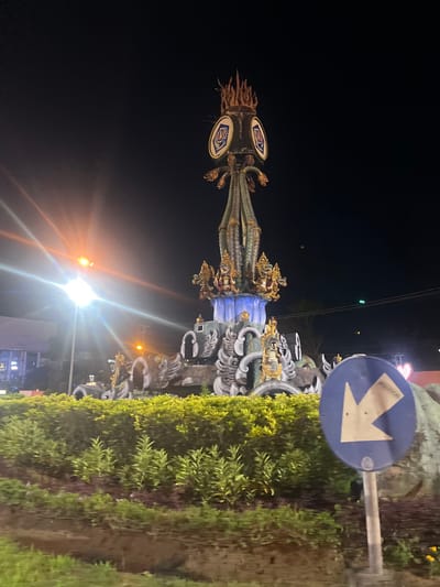 The image shows a large monument in Denpasar, Indonesia, at night. The monument is elaborately decorated with intricate carvings and appears to be made of stone and metal. It features multiple tiers, with what look like dragon-like sculptures and golden accents. At the very top, there is a fiery element. The monument is surrounded by lush green bushes and plants. A street light with bright flares illuminates the scene from the left, casting light on the monument and the surrounding area. In the foreground, to the right, a blue circular traffic sign with a white arrow pointing left and the word "POLITICAL" in small text is visible. The background is dark, indicating nighttime, with some faint lights from distant buildings and power lines. The overall impression is of a significant cultural landmark in a city setting.