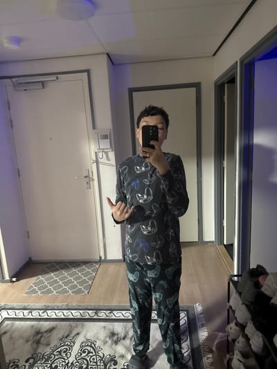 An individual is depicted in a full-body selfie taken in a mirror within an indoor residential setting, likely an apartment hallway or entryway in Voorburg, Netherlands. The subject, with short, dark, curly hair, holds a dark-colored smartphone to obscure their face. They wear a dark-colored, two-piece pajama or loungewear set featuring an all-over pattern of video game controllers in white and blue accents, along with dark grey or black slip-on slippers. A ring is present on the subject's left ring finger. Their right hand (viewer's left) performs a "shaka" or "hang loose" gesture.
The background reflected in the mirror reveals a white interior door with a dark frame, equipped with a silver-colored handle, a peephole, and an overhead hydraulic door closer. Adjacent to this door, a white intercom or entry system panel is mounted on the wall. The flooring consists of light-colored wood or laminate, with a smaller, grey and white patterned rug placed in front of the white door. A larger, dark patterned rug with ornate designs is visible in the foreground reflection. To the subject's right, another doorway with a dark frame leads into an unlit or darker interior space. On the far right, a dark metal multi-tiered shoe rack holds several pairs of shoes. The ceiling is white with a round, flush-mounted light fixture, and blue ambient light reflections are visible on the ceiling and walls.