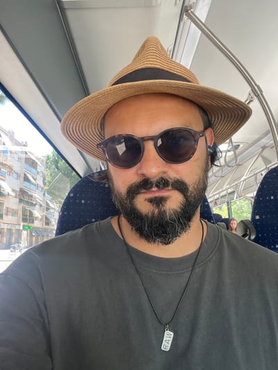 The image shows a close-up, low-angle shot of a person wearing a straw hat and sunglasses, with a beard. The person is wearing a dark grey t-shirt and a necklace with a rectangular pendant that reads "RAW". The scene appears to be on a mode of public transportation, possibly a train or bus, with blue patterned seats and metal handrails visible. Through the window, a modern building with many windows and balconies can be seen, suggesting an urban or semi-urban setting. The lighting suggests it is daytime, and the weather appears to be clear. The overall mood is casual and relaxed, with the subject possibly taking a selfie. The building outside has some visible signage, including "SOLARISTAS" and "SOLARISTAS S.L.".