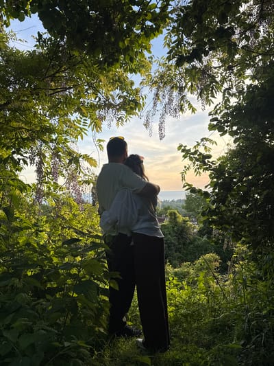 A couple is embracing in Tsanbaikwara, Abkhazia, with a sunset in the background. They are standing amidst lush greenery, with trees and foliage framing the view. The person on the left is wearing sunglasses, a white top, and dark trousers. The other person has long hair and is wearing a white top. They appear to be hugging and looking up toward the sky.