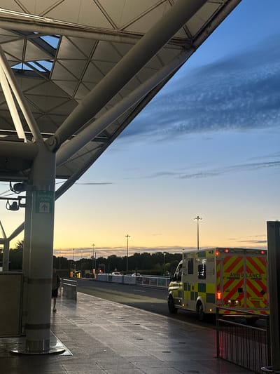 An exterior shot captures a pavement at what appears to be an airport terminal in Uttlesford, United Kingdom, during dusk. The sky displays hues of blue and orange with wispy clouds. A large, modern roof structure with exposed beams and triangular panels extends over the walkway. To the left, a "Smoking Area" sign with an upward arrow is affixed to a pillar. A person in casual clothing, a t-shirt and shorts, stands near the pillar. An ambulance, identified by the text "AMBULANCE" on its rear, is parked on the road to the right, its lights illuminated. The road is bordered by railings and a row of trees and streetlights are visible in the background. The ground is paved with large tiles, and the lighting suggests it may be damp.