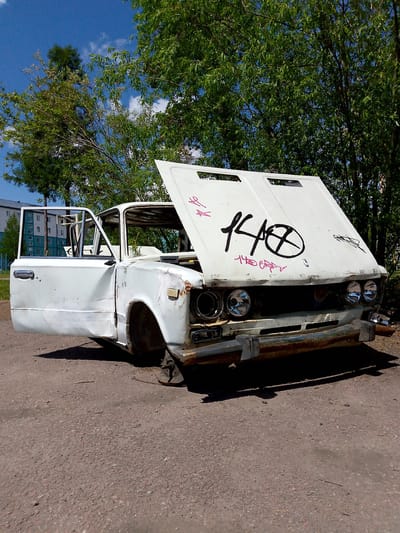 The image depicts a dilapidated white car abandoned on a dirt road in Neryungri, Russia. The car, likely a Soviet-era Lada based on its design, is heavily damaged and appears to be stripped of its wheels and other components. The driver's side door is open, revealing an empty interior. Graffiti adorns the hood of the car, with prominent black lettering and a circular symbol. The background features lush green trees and a bright blue sky with scattered clouds. In the distance, a modern apartment building is visible, suggesting an urban or semi-urban setting within Neryungri. The scene conveys a sense of neglect and decay, juxtaposed against the natural beauty of the surrounding environment.
