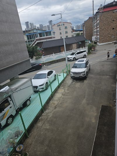 This is an eye-level shot of a street scene in Jincheon-eup, South Korea. In the foreground, a white truck with a silver cargo box is parked to the left, its wheels visible. Adjacent to the truck is a green fence bordering a teal-colored surface. Several white cars are parked on the street, including a white sedan with a license plate reading "0383". Further down the street, a white SUV with a license plate "650 9053" is parked. A young girl in a black skirt and white top is walking away from the viewer on the right side of the street, near some blue plastic bags. Behind the parked cars, residential buildings with brick and stone facades, as well as modern apartment buildings, are visible. The sky is overcast with scattered clouds.