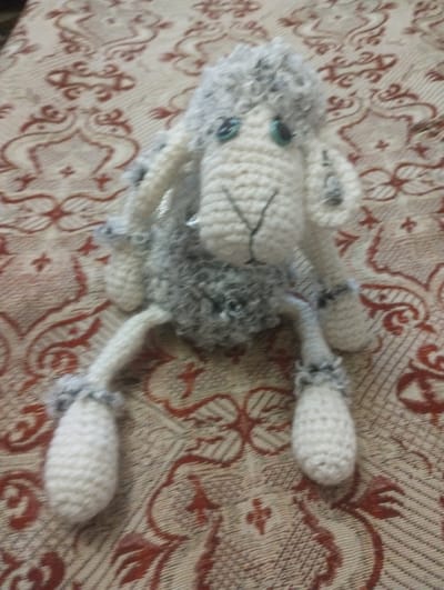 The image shows a handmade, crocheted toy rabbit lying on a patterned fabric surface. The rabbit is primarily off-white with a grey, fluffy section around its neck and head. It has two blue button eyes and a stitched mouth. The toy's limbs are elongated and also crocheted in the off-white yarn. The fabric underneath the toy has a repeating pattern of stylized red and beige floral or paisley designs. The lighting in the image is soft and diffused, suggesting it was taken indoors during the daytime, possibly in a home setting in Bilany, Ukraine. No text is visible in the image. The overall impression is of a cozy, domestic scene, with the toy rabbit appearing as the central focus. The camera angle is from above, looking down at the toy.