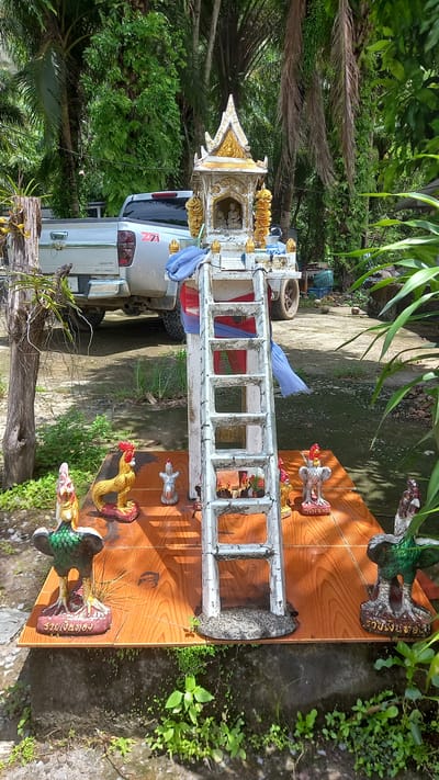 The image shows a spirit house in Thap Put, Thailand. The spirit house is a white, multi-tiered structure with ornate gold detailing and a ladder-like attachment. Colorful cloths are draped over the ladder. The spirit house sits on a wooden platform adorned with figurines of roosters and other animals. The platform rests on a concrete surface, surrounded by greenery. A silver pickup truck is parked in the background, partially obscured by trees. The environment appears to be a residential or semi-rural setting with vegetation and a paved area.