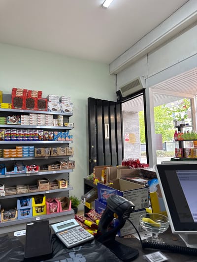 The image depicts the interior of a small shop or convenience store in Lurgan, United Kingdom. The scene is taken from behind the checkout counter. There's an open doorway at the back of the store. The shelves are stocked with a variety of packaged goods, including biscuits and chocolate. A barcode scanner and a calculator are visible on the counter, along with a digital point-of-sale system. There are no people visible in the image. A "no smoking" sign is posted near the door.