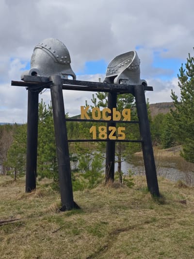 A sign in Косья, Russia, features two metal helmets atop a black frame. The frame displays the word "Косья" in yellow lettering above the number "1825." The sign stands on a grassy hill overlooking a body of water, with a backdrop of trees and a cloudy sky.