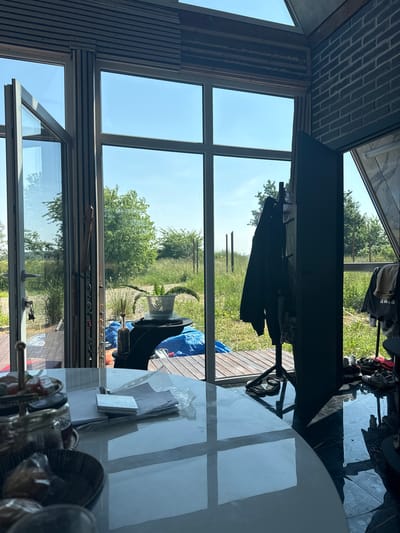 This media file presents an indoor view looking out through large windows and a glass door, offering a glimpse of an outdoor scene. The interior features a modern, minimalist design with exposed brick on the ceiling and wooden paneling on the walls. The foreground is dominated by a reflective surface, likely a table or countertop, showing reflections of the surroundings and some objects, including what appear to be decorative items or containers.
Outside, the scene depicts a grassy, overgrown area with trees and foliage under a clear blue sky. The presence of a clothes rack with several garments hanging on it suggests laundry is being air-dried or simply stored outdoors. A blue tarp is visible, possibly covering something or serving as a ground cover. The lighting indicates it is daytime, likely late morning or early afternoon, with bright sunlight casting shadows.
The overall atmosphere is one of a casual, perhaps rural, dwelling with an emphasis on natural light and an connection to the outdoors. The image is oriented vertically, making it appear as if it was taken while standing or from a low angle, looking upwards. The lack of people suggests a solitary or unoccupied moment. The location context of Vol'noe, Russia, hints at a potentially temperate climate, consistent with the green vegetation and clear skies.
