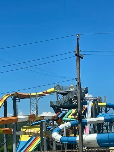 The image shows a large water park with multiple slides of various colors and shapes. The slides are made of plastic and metal, and they are arranged in a complex structure with walkways and stairs. The water park is located outdoors, and the sky is blue with some clouds. The image also shows a utility pole with power lines in the foreground. The water park is likely a popular tourist attraction in Lermontovo, Russia.