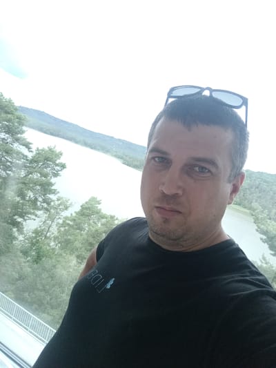 A man is taking a selfie in front of a large body of water and a hillside covered in trees. The man is wearing a black t-shirt and has sunglasses on his head. The body of water appears to be Lake Macha in Doksy, Czechia. The weather is overcast.