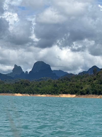 The image depicts a wide view of Cheow Lan Lake in Khao Sok National Park, near Ban Chiao Lan, Thailand. In the foreground, a body of turquoise water is ruffled by gentle waves, suggesting a boat or other vessel is present, as indicated by a faint shadow on the left side. The middle ground features a tree-lined shore with a band of dry, reddish-brown earth. Beyond the trees, the landscape rises dramatically with several large, dark green, forest-covered mountains dominating the background. The sky is filled with dramatic, grey and white clouds, hinting at overcast or potentially stormy weather. The overall scene is one of natural, rugged beauty.