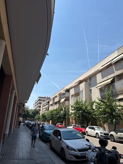 This is a vertical, wide-angle, eye-level shot of a street in Vilanova i la Geltrú, Spain, on a sunny day. The sky is a clear blue with two distinct white contrails. On the left side of the frame, a large, light-colored building with balconies and horizontal blinds dominates the upper portion. Its reflection is visible on the dark, textured facade of a building in the foreground.
The street is lined with parked cars and scooters. A white Peugeot car with license plate "8588 NFR" is prominently parked towards the right. Further down the street, a white Peugeot car with license plate "8588" is visible. A woman in a blue top and denim shorts is walking on the sidewalk, carrying a white bag. Another person in a black outfit is walking further down the street. Trees with lush green foliage are scattered along the sidewalk, casting shadows. The buildings on the right are multi-story apartment complexes with balconies. The time of day appears to be late morning or early afternoon, suggested by the bright sunlight and the length of the shadows. The overall mood is calm and ordinary, depicting a typical day in a European town.
