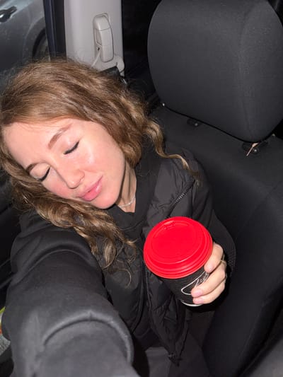 A young woman with curly brown hair is reclining in the passenger seat of a car. She is wearing a black puffer jacket and holding a black coffee cup with a red lid in her right hand. The interior of the car is mostly black. A car seat and the interior of the car are visible. The location in Krilovo, Russia is not ascertainable from the image.