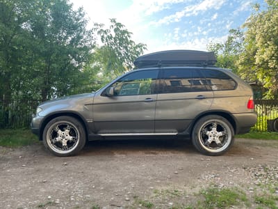 The image depicts a grey BMW X5 SUV parked on a gravel surface, possibly in the city of Kalnciems, Latvia, given the context. The vehicle has a dark-colored roof box mounted on its roof. The surrounding environment features lush green trees, some of which are in bloom with white flowers, and a fence in the background. The sky is visible with scattered white clouds. The focus is on the side profile of the car, showcasing its custom rims and a side step. The lighting suggests it is daytime, likely late afternoon or early morning, given the soft shadows. There are no people visible in the image.