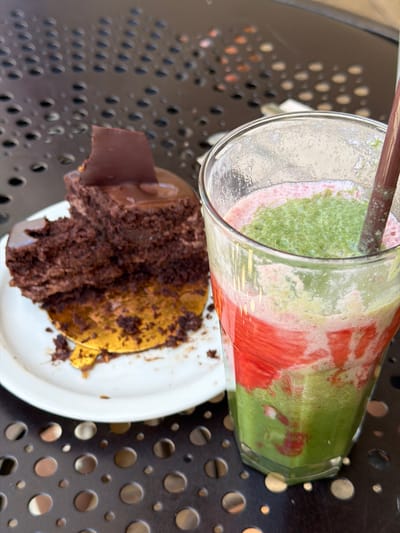 A slice of chocolate cake sits on a white plate next to a glass of pink and green layered smoothie, all on a black metal table with a circular pattern. The cake is dark brown with a chocolate shard on top and appears to be a layered mousse cake. Crumbs are scattered on the plate. The smoothie has a thick, frothy texture and is being stirred by a brown straw. The scene is lit from above and to the right, casting shadows. The location is not specified in the image, but the table appears to be outdoors, possibly at a cafe or restaurant.
