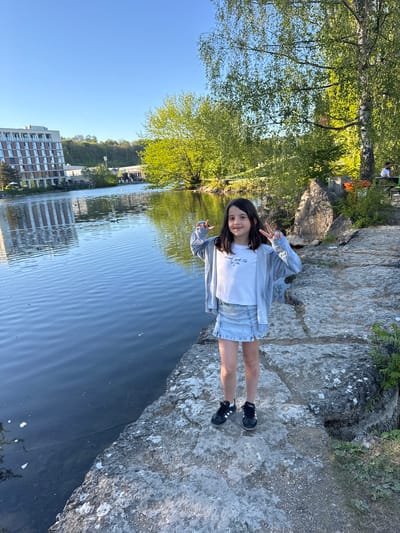 This image captures a young girl posing cheerfully outdoors on a bright, sunny day in what appears to be Ulm, Germany. The girl, with dark hair, stands on a rugged, light-colored rocky bank adjacent to a calm body of water, likely a river or canal. She wears a white t-shirt, a light gray zippered hoodie, a light blue denim skirt, and black Adidas sneakers with white stripes. With a bright smile, she holds up both hands, making peace signs, conveying a happy and relaxed mood. A small scrape is visible on her left knee.
Her white t-shirt features discernible text that reads "Mariani Sports Club" with "New York City" written underneath.
The background showcases lush, vibrant green trees lining the opposite side of the water, with their reflections shimmering on the surface. Beyond the trees, the clear blue sky dominates the upper portion of the scene. In the further distance, several modern, multi-story buildings are visible, indicating an urban setting bordering natural landscapes. The bright sunlight illuminates the entire scene, highlighting the textures of the rocks and the subtle ripples on the water. The overall impression is that of a pleasant outdoor moment, combining natural elements with urban architecture.