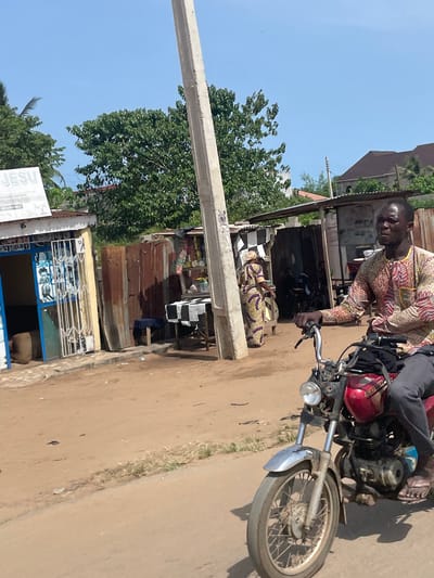The image depicts a man on a motorcycle on a street in Ikoga, Nigeria. The man is wearing a patterned long-sleeved shirt. He is seated on a red motorcycle with a rusted front fender. Behind him, a woman is sitting on a low chair in front of a building with a corrugated metal wall. The building appears to be a shop or business. A utility pole runs vertically along the left side of the frame. In the background, other buildings and trees are visible under a clear sky. The road is unpaved.