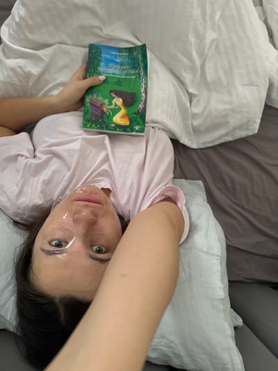 A female individual lies on a bed in Trekhgorka, Russia. She is positioned with her head on a white pillow, facing upwards, and wears a light pink short-sleeved top. A translucent, gel-like face mask is applied to her face, with her eyes open and directed towards the camera. Her right arm is raised, holding a book with a green cover against her chest. The book features an illustration of a woman with long hair near a beehive in a natural setting and bears the Russian title "Сундучок волшебных историй" (Chest of Magic Stories) and "Терапевтические сказки" (Therapeutic Tales) by "Наталья Веда." White bed linens surround her upper body, and a dark grey sheet or blanket covers the lower portion of the bed.