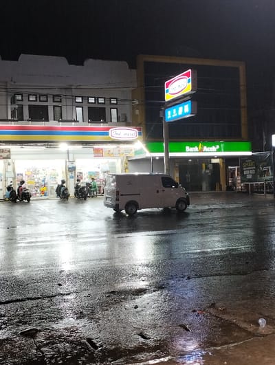 This image captures a nighttime street scene in Jeunieb, Indonesia. The ground is a dark, wet asphalt surface, highly reflective of the artificial light sources, indicating recent rain or standing water.

On the left, a two-story building houses an illuminated "Indomaret" convenience store on its ground floor, identified by its distinctive red, blue, and yellow logo and bright interior lighting. The upper floor of this building is white with multiple dark, rectangular windows.

Adjacent to Indomaret on the right, a "Bank Aceh" branch is visible, featuring a green facade with white lettering. A tall pole sign stands between Indomaret and Bank Aceh, displaying the "Indomaret" logo at the top and a blue sign with white icons below. Further to the right, a darker building is partially framed by a yellow structure.

A light-colored, possibly white or light grey, commercial van is positioned on the wet street in the mid-ground, facing towards the right side of the frame. Several motorcycles are parked or idling near the entrance of the Indomaret store, with at least four discernible. Multiple individuals are present within the illuminated Indomaret entrance area, some appearing to be associated with the motorcycles, others standing.

The scene is dominated by artificial lighting from the commercial establishments and signage, which creates strong reflections on the wet road. The sky above is dark. Power lines are visible traversing above the buildings. In the foreground, a white plastic cup and minor debris are discernible on the wet road surface. An advertisement, partially visible on the far right, includes the word "SMART."