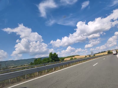 The image shows a bright, sunny day with a blue sky filled with fluffy white clouds. The perspective is from a moving vehicle on a paved road, indicated by the presence of a metal guardrail and dashed white lane markers. The road curves to the right in the distance, where a white truck with "EKO" visible on the back is traveling.
To the left of the road, there is a hilly landscape. Green trees dot the foreground and midground, with some dry, dead trees also present. Beyond the trees, rolling hills are covered in patches of green and golden fields, suggesting farmland. The distant hills are a deep green.
The weather appears to be warm and clear, typical of a summer day. The lighting suggests it is daytime, likely late morning or afternoon, given the bright sunlight and distinct shadows. The overall mood is one of travel and open scenery. The location is identified as Dzhurovo, Bulgaria. There are no people visible in the image.