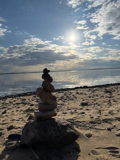The image shows a stack of rocks on a beach, with the sea and sky in the background. The sky is partly cloudy with a bright sun. The beach is sandy with some small rocks and debris. The stack of rocks is composed of different sizes and shapes. The location is not specified in the image. I cannot place this image in Yasenevo, Russia.