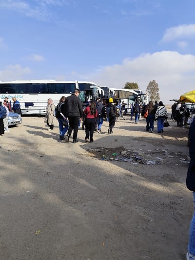 An outdoor scene in Mankat al-Hatab, Syria, depicts a bus station or large transit area under a partly cloudy blue sky. In the background, a row of at least three white passenger buses with dark tinted windows and blue "Misk Travel Tours" branding are parked. The leftmost bus has a silver sedan parked in front of its passenger side. A crowd of approximately twenty-five individuals, primarily adults and young adults of varying ages and genders, are visible in the mid-ground and foreground, walking across the dusty, uneven ground which appears to be a mix of dirt and degraded asphalt. Many individuals carry backpacks or bags; notable attire includes a black puffer jacket, a red long-sleeved top, a yellow beanie, and a black and white patterned jacket. One woman wears a beige hijab and coat. On the far right, a yellow market canopy or umbrella is partially visible. The ground in the foreground displays scattered litter, including discarded papers, plastic, and cigarette butts, with a concentrated pile of debris visible in the middle-right. Two sparse trees are present in the distant mid-ground on the right. Shadows are cast on the ground from the people and unseen objects to the left, indicating daylight conditions.