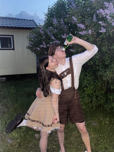 Two people, a male and a female, are lying on grass, presumably in Пальники, Russia. The male is wearing a traditional Bavarian outfit with brown lederhosen, a white shirt, and suspenders, and is drinking from a green bottle. The female is wearing a short beige dress with embroidered trim and white socks with black shoes. In the background is a lilac bush and a house with a brown roof and yellow siding. The lighting suggests it is dusk or evening.