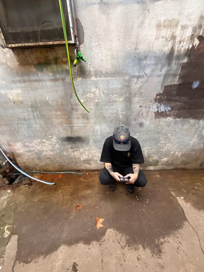 The image is a top-down view of a person crouching on a weathered, concrete surface, possibly a courtyard or alleyway. The person is wearing dark clothing, a hat, and appears to be holding a smartphone. The surface is stained and uneven, with a window or grate visible in the bottom left corner. A yellow and green garden hose is attached to a spigot nearby. The environment seems neglected, with debris and discoloration adding to the unkempt atmosphere. The photo location is Villa Bernardo de Monteagudo, Argentina.