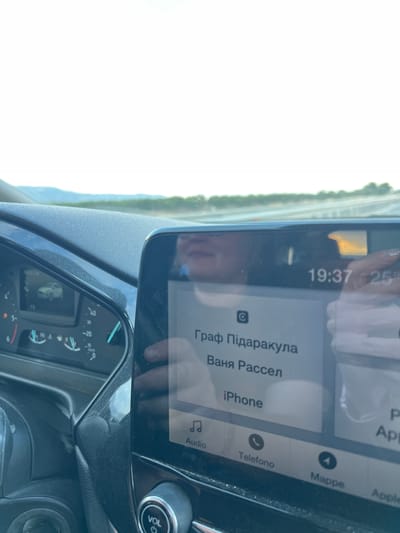 The image shows the interior of a car, likely a Ford Focus, as indicated by the dashboard display. The car is in motion, as evidenced by the blur of the landscape outside and the readings on the speedometer, which shows a speed of approximately 20 km/h. On the central infotainment screen, the time is displayed as 19:37 and the temperature as 25 degrees. The screen also shows a contact name, "Граф Під'яракула," and a sub-name "Ваня Рассел," suggesting a phone contact or a music playlist. Below this, "iPhone" is displayed, indicating the connected device. The bottom of the screen shows icons for "Audio," "Telefono," "Mappe," and "Apple." The scene is captured from the perspective of the driver or front passenger, looking towards the dashboard and the infotainment system. The exterior landscape appears to be a rural or semi-rural area with rolling hills and vegetation under a bright sky. There is no direct evidence to place this specific image in Manfredonia, Italy.