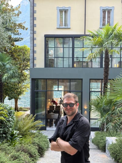 A smiling man in a black collared shirt and sunglasses stands with his arms crossed in front of a modern building with large glass windows and palm trees. The man is positioned on a paved walkway lined with lush green bushes. In the background, the building features a pale yellow facade with a darker grey lower section and a lake with mountains visible behind it. The interior of the building through the glass shows a dining area with tables and chairs, and two figures seated. The scene appears to be a residential property in Pognana Lario, Italy, likely a villa given the architectural style and lakeside setting.