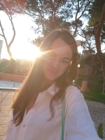 A woman with long brown hair and fair skin is taking a selfie outdoors. She is wearing a white collared shirt and a light green strap is visible across her chest. The sun is shining brightly, causing lens flare and obscuring parts of the background. In the background, there are trees, some buildings, and a paved walkway. The scene appears to be in a park or public outdoor area within the city of El Campello, Spain, likely near the coast given the natural light and vegetation. The time of day is likely late afternoon or early evening due to the low angle of the sun and the warm light.