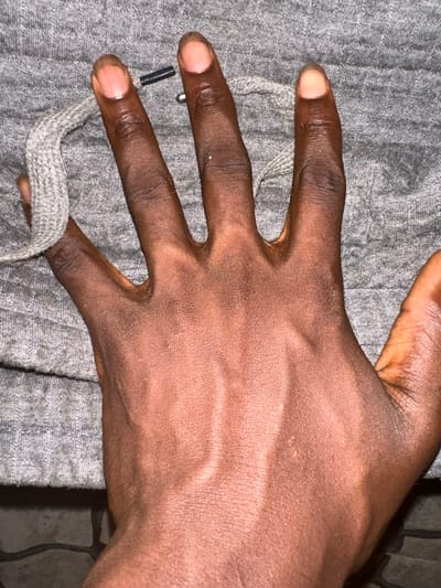 A close-up shot reveals a hand resting on a gray surface. The hand's palm is visible with its veins slightly raised, suggesting a strong grip. The skin tone is dark brown. Two fingers cradle a braided gray cord, possibly a drawstring from clothing. The background appears to be fabric of the same gray color, adding a textural contrast to the smooth skin. The lighting is soft, accentuating the hand's contours.