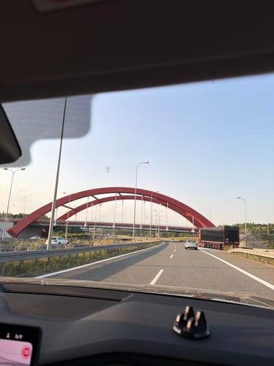 The image is taken from the interior of a vehicle on a highway. In the distance, a large red arch bridge spans across the highway. Several vehicles, including cars and a semi-trailer truck, are visible on the road. Streetlights line the highway. The sky is clear and blue. The scene appears to be set in the city of Przyszowice, Poland.