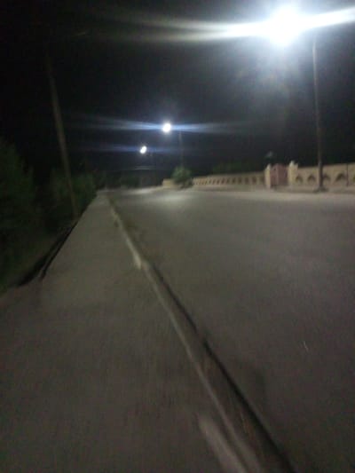The image depicts a nighttime scene on a street in Shar-Bakr, Uzbekistan. A paved road occupies the foreground and middle ground, with a concrete barrier or sidewalk to the left. Streetlights illuminate the scene, with one bright light source dominating the upper right portion of the frame. In the background, a low wall with arched decorative elements runs along the side of the road, suggesting a property boundary or architectural feature. Vegetation is visible to the left and in the distance. The overall impression is one of an urban or semi-urban environment at night.