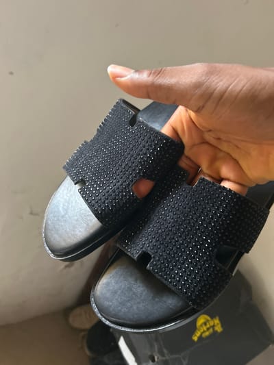 The image captures a person's hand holding a pair of black slide sandals, presented against a light-colored, plain background in an indoor setting. The hand, with medium to dark brown skin, gently grasps the sandals, showcasing their design.

The sandals feature wide straps heavily embellished with numerous small, sparkling elements, creating a textured and reflective surface. The footbeds are dark, possibly black or dark grey, and the soles are black. In the upper right background, a black box with the yellow "Dr. Martens" logo is partially visible, suggesting brand context or packaging. Another pair of shoes or sandals is indistinctly seen next to the box.

The primary activity depicted is the presentation or examination of the black embellished sandals. The lighting is even, typical of an indoor environment, providing no specific cues regarding the time of day or outdoor weather. No emotions are discernible from the partial view of the hand. The scene is neutral, focusing primarily on the footwear. The location context is Ashipa, Nigeria.